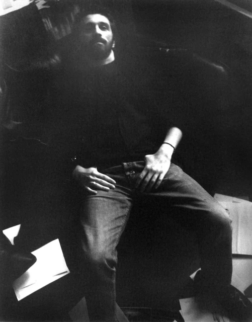 Monochrome portrait of a bearded adult male seated with his hands on his lap wearing casual clothing in a dimly lit room.