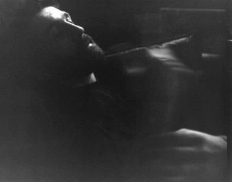 Black and white low-key portrait of a bearded man leaning back in deep shadow with light catching his facial profile.