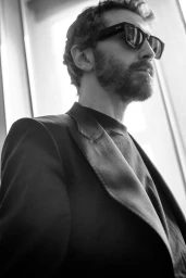 Low-angle black and white portrait of a bearded man wearing dark sunglasses and a tailored black jacket by a bright window.