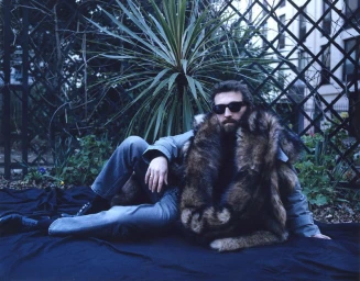 A bearded man wearing dark sunglasses and a voluminous fur coat reclines on dark fabric in front of garden foliage and a trellis.