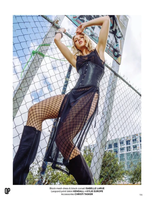 Fashion editorial shot of a slender adult female model wearing a black mesh dress and thigh-high boots posing by a chain-link fence outdoors.