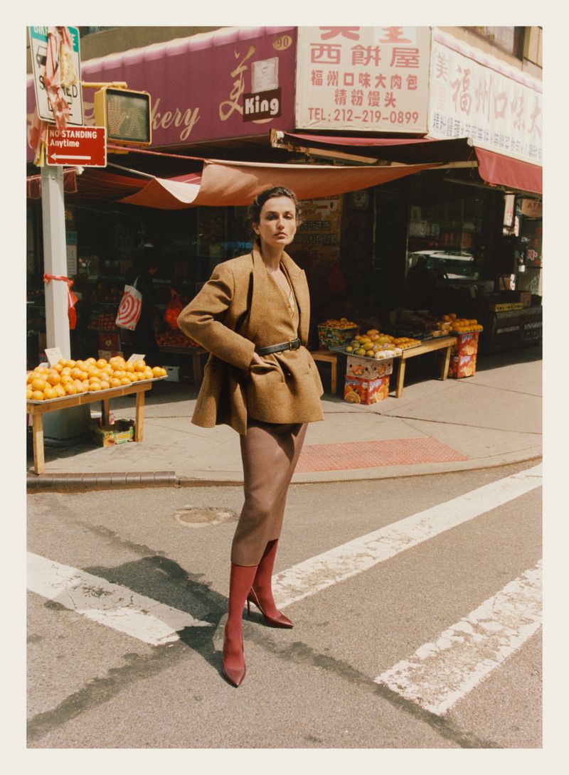 Adult woman in a tan jacket and pencil skirt poses on a crosswalk in front of a bakery with orange fruit stands outside.