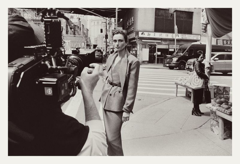 Fashion photoshoot on a city street with a model posing in a suit, camera crew visible, and pedestrians in the background.