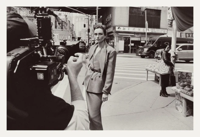 Fashion photoshoot on a city street with a model posing in a suit, camera crew visible, and pedestrians in the background.