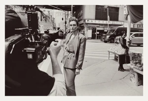 Model standing on a city street in a tailored blazer and skirt, filmed by a camera crew in the foreground.