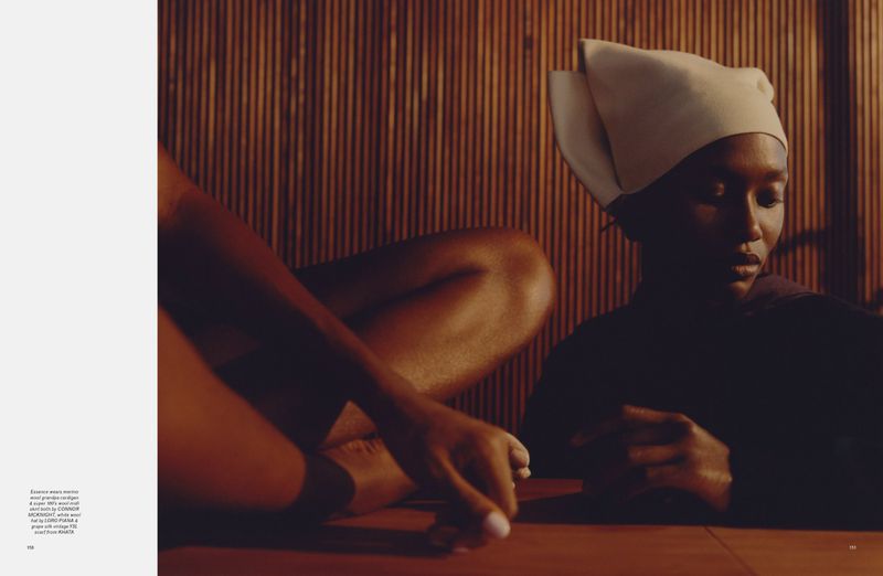 Black model wearing an off-white sculptural headpiece seated in a warm, dimly lit wooden interior with another figure.