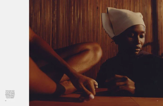 Black model wearing an off-white sculptural headpiece seated in a warm, dimly lit wooden interior with another figure.