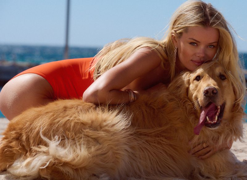 Blonde woman in swimsuit laying on a golden retriever dog at the beach with a blue ocean backdrop during a sunny day.