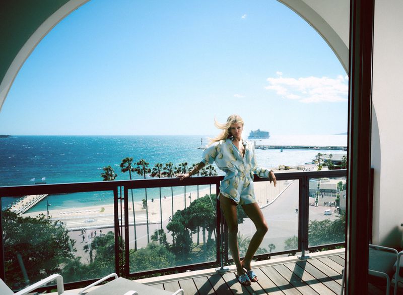 Fashion shot of a blond woman posing on a balcony overlooking a beach with palm trees, turquoise water, and a cruise ship in the distance.