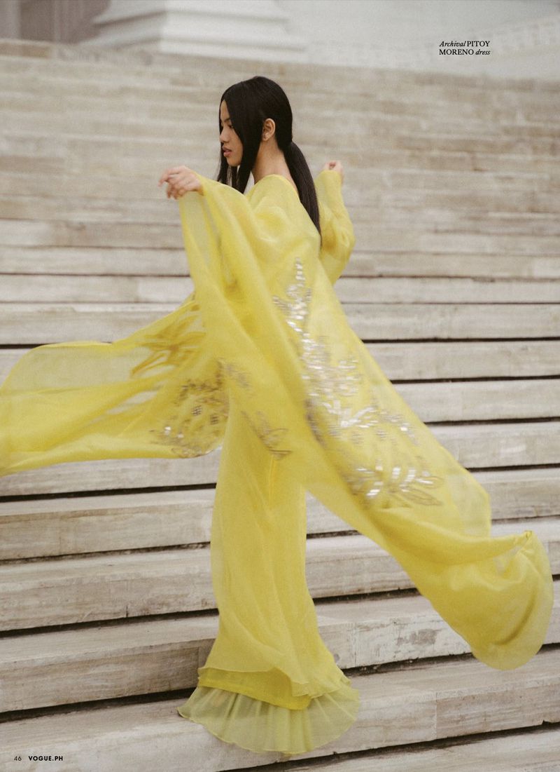 Elegant woman in a flowing yellow gown poses on stone steps with column detail in the background, fashion editorial, vogue.