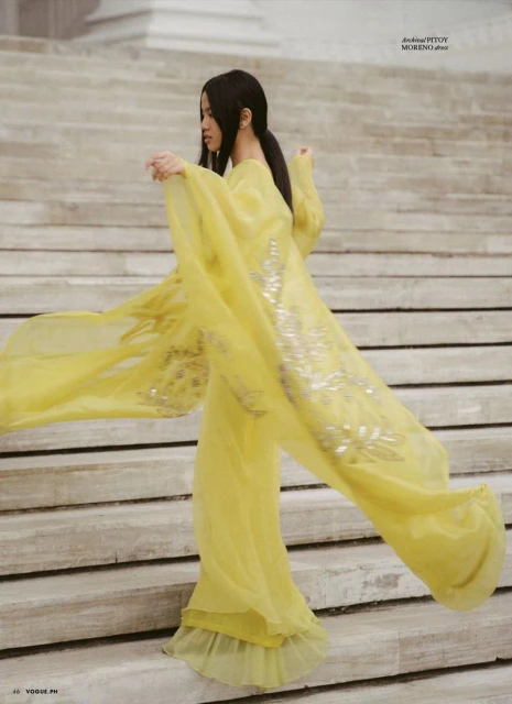 Elegant woman in a flowing yellow gown poses on stone steps with column detail in the background, fashion editorial, vogue.
