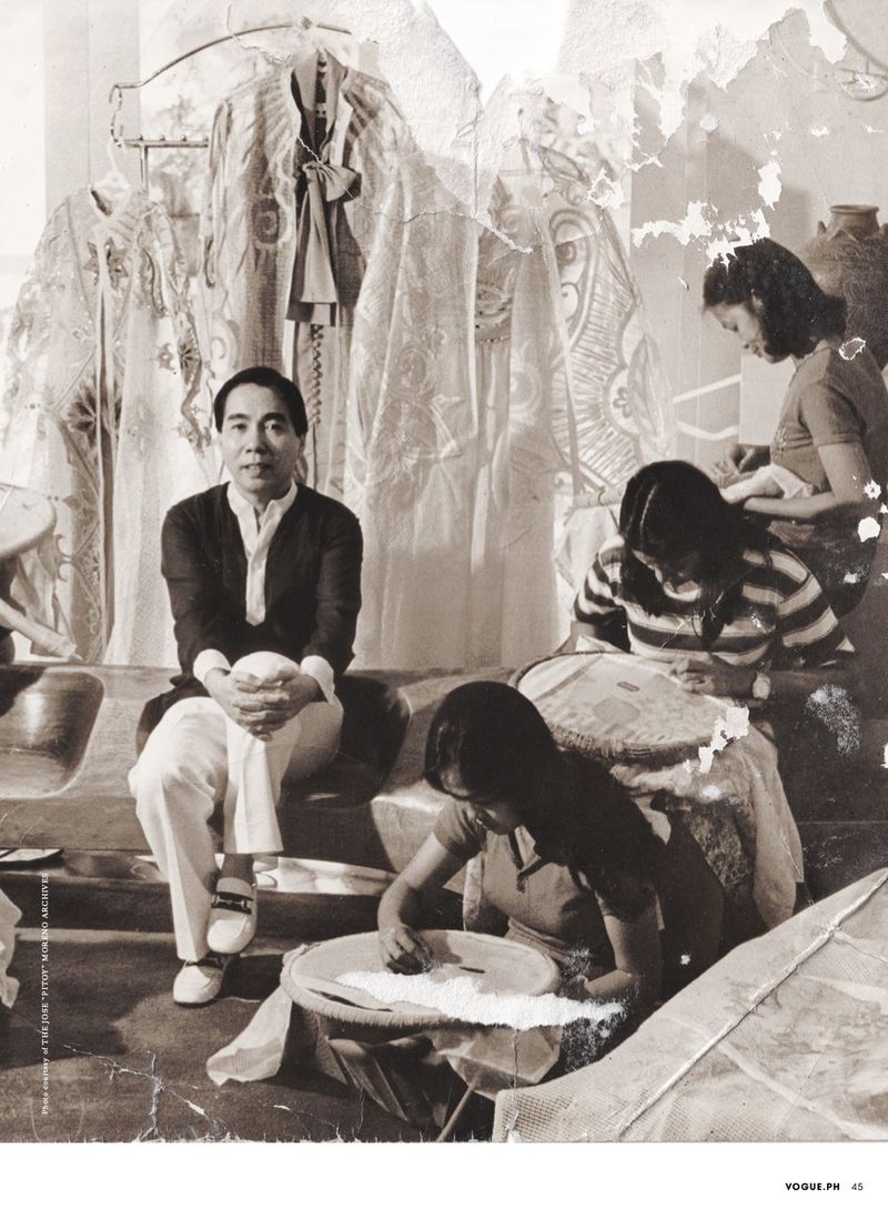 Monochrome photograph of an Asian man sitting on steps in front of women hand-sewing, with textiles hanging in the background.