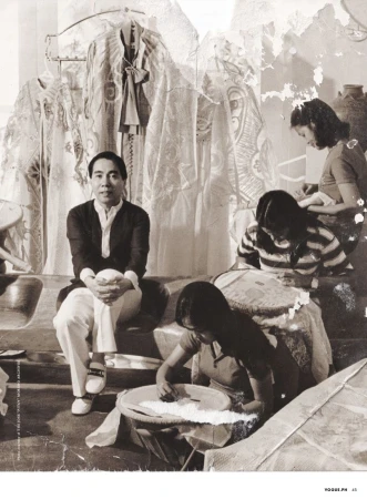 Monochrome photograph of an Asian man sitting on steps in front of women hand-sewing, with textiles hanging in the background.