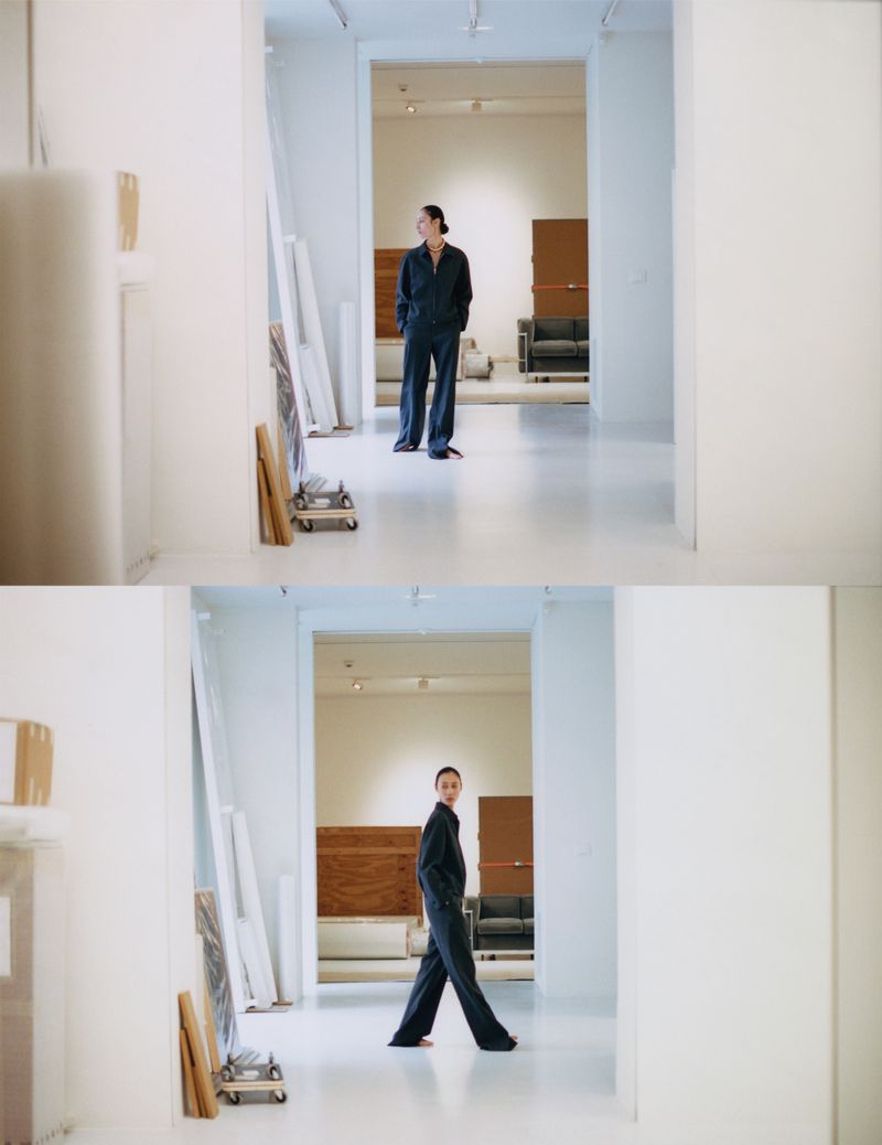 Woman in dark jumpsuit stands in a bright white art gallery, surrounded by canvases and art supplies, creating an atmosphere of creativity.