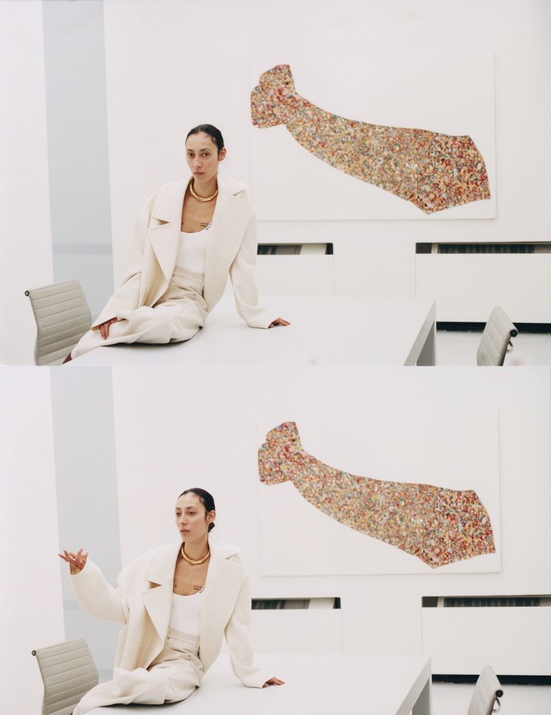 Stylish adult woman poses on a white table in an indoor setting wearing a cream-colored suit with modern art in the background.