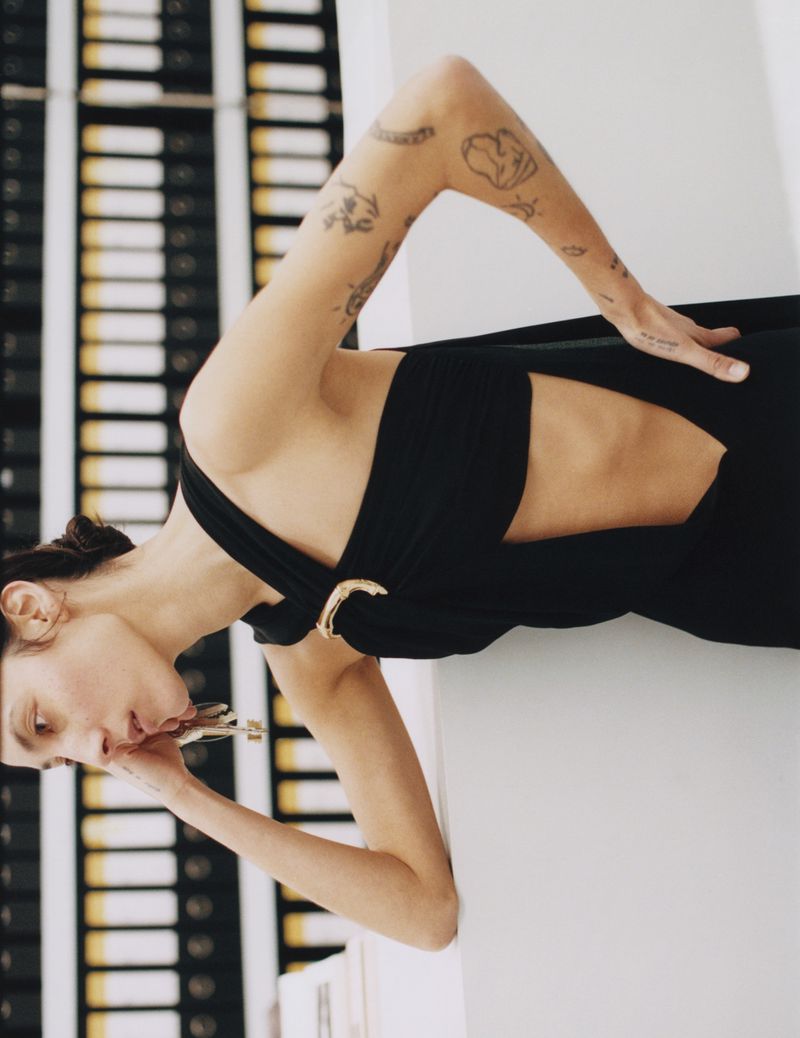Adult woman poses sideways in black dress with cut-out and gold clasp detail, in an indoor setting with neutral wall and shelving.