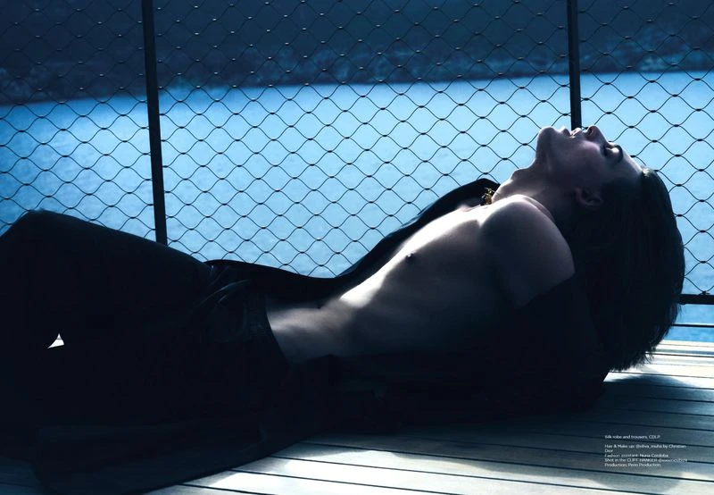 Man lies on a wooden deck near a railing with a blue water background, wearing black clothing and looking upward in a relaxed pose.