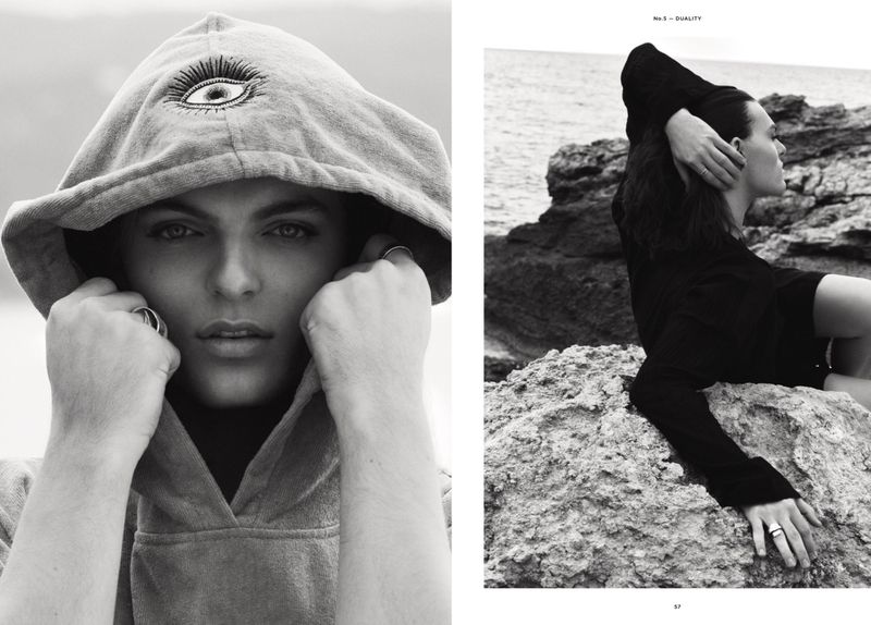 Monochrome portrait of a person in a hoodie with a graphic eye and another person in a sweater on a rocky coast by the ocean.