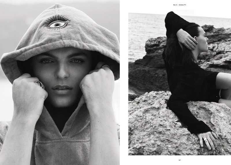 Monochrome portrait of a person in a hoodie with a graphic eye and another person in a sweater on a rocky coast by the ocean.