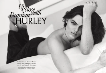 Monochrome portrait of Damian Hurley lying on a white bed with long dark hair, wearing dark trousers and a necklace.