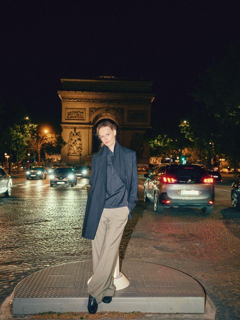 Adult woman poses in a dark coat and khaki pants on a city street in Paris with the Arc de Triomphe in the background at night.
