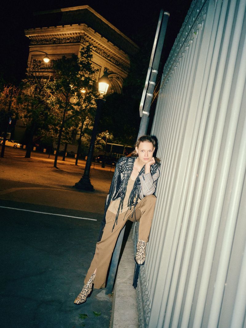 A young woman in patterned jacket and animal-print boots poses on a street at night, with the Arc de Triomphe in the background.