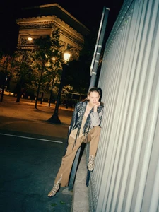 Cosima Fritz wearing beige trousers, a fringed shawl, and snakeskin boots leaning against a street pole near the Arc de Triomphe at night.