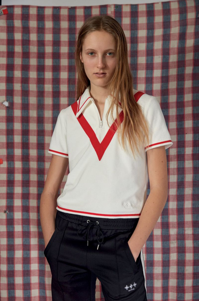 Young adult female posing in a white t-shirt with a red V-neck design and black sweatpants, indoors against a plaid backdrop.