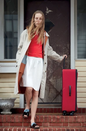 Young adult woman with luggage descends steps of a house in a light trench coat, red shirt, and white skirt on a cloudy day.