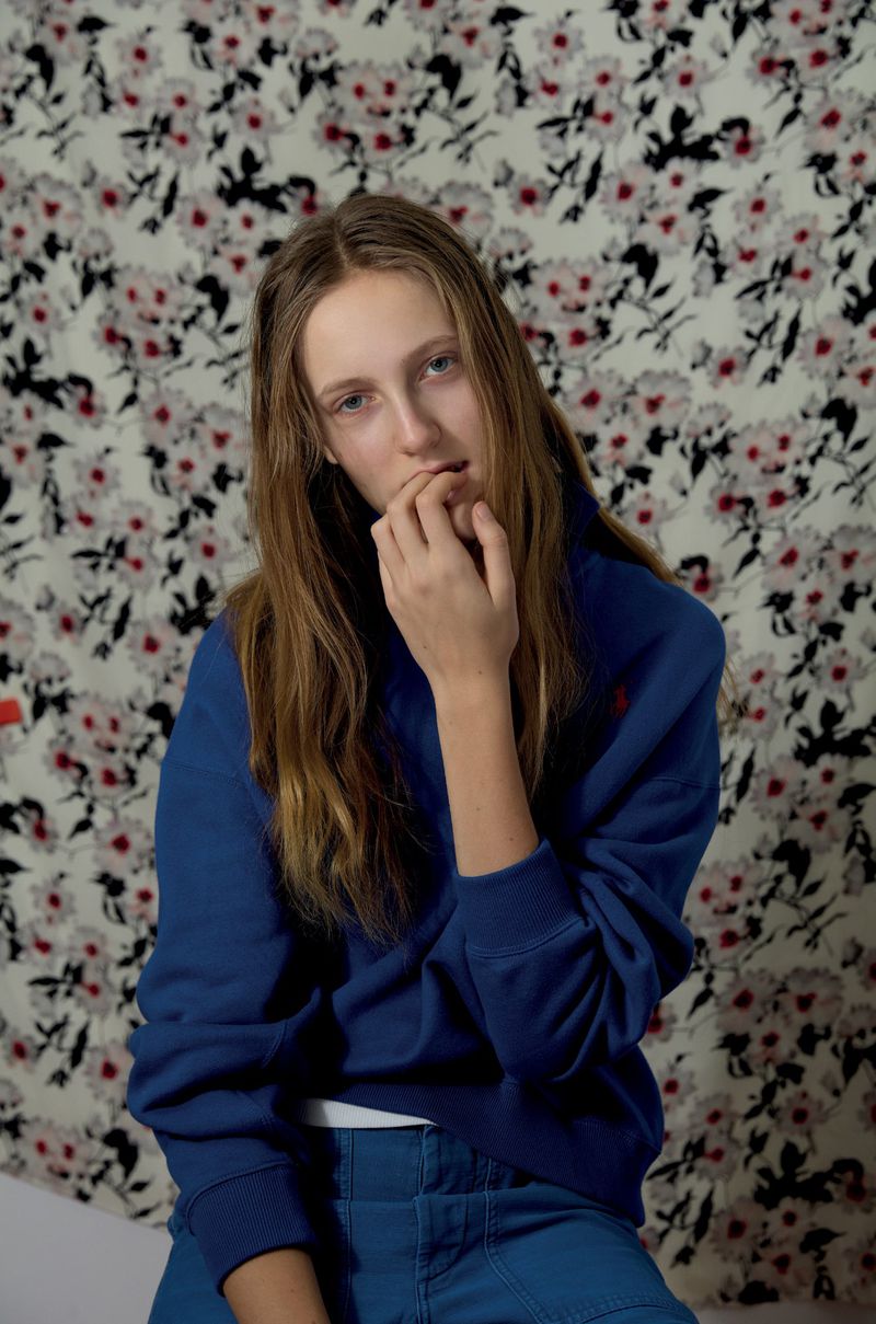Young woman with long hair poses indoors against a floral wall, wearing a blue Ralph Lauren sweatshirt and blue pants, looking at the camera.