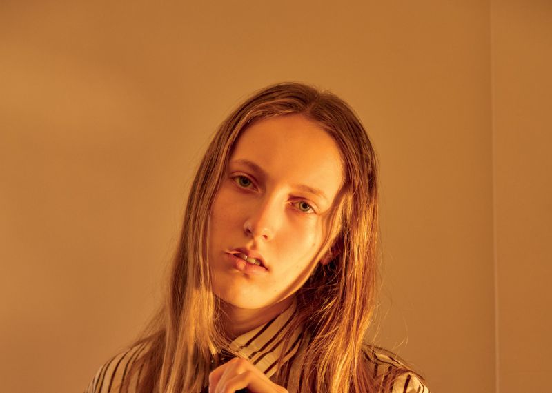Close-up of a young adult woman with long blonde hair posing in front of a warm toned wall, wearing a striped shirt and tie.