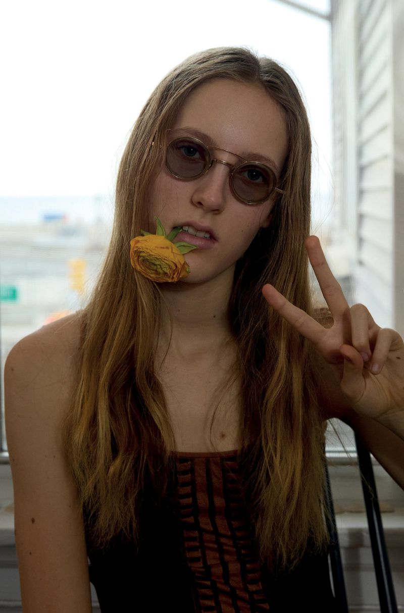 A woman wearing round tinted sunglasses with a yellow flower in her mouth, gesturing a peace sign indoors by a window.