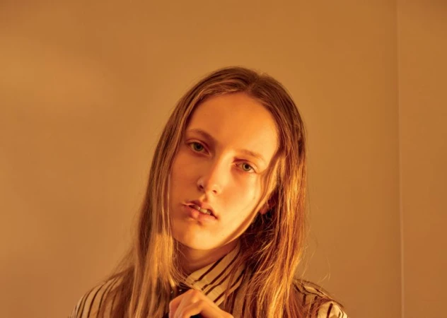 Close-up portrait of a person with long brown hair wearing a striped shirt against a plain warm-toned wall