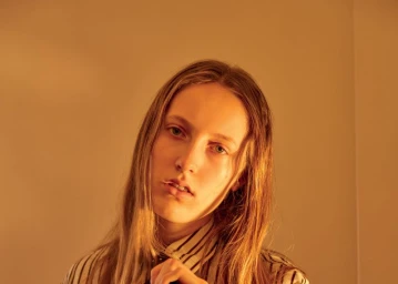 Close-up portrait of a person with long brown hair wearing a striped shirt against a plain warm-toned wall