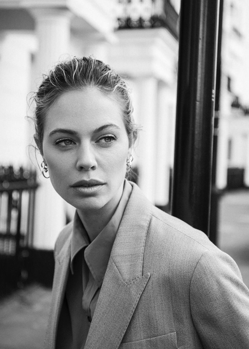 Black and white portrait of an adult woman in a blazer and collared shirt outdoors with a blurry background of buildings and street lamp.