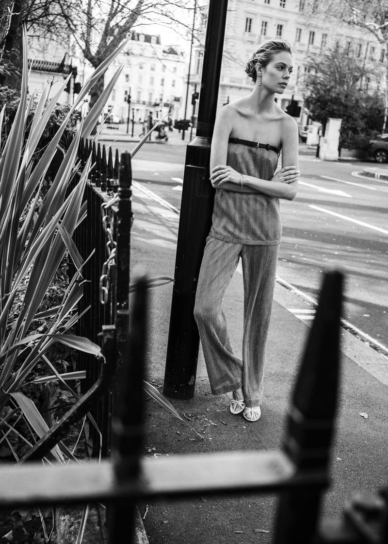 Black and white portrait of a fair-skinned woman wearing pants and a strapless top standing on a city street with arms crossed.