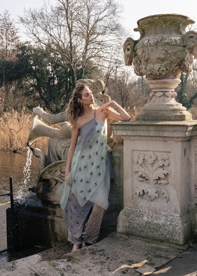 A woman in an asymmetrical dress stands by a decorative fountain with an urn in a park setting with trees and natural light.