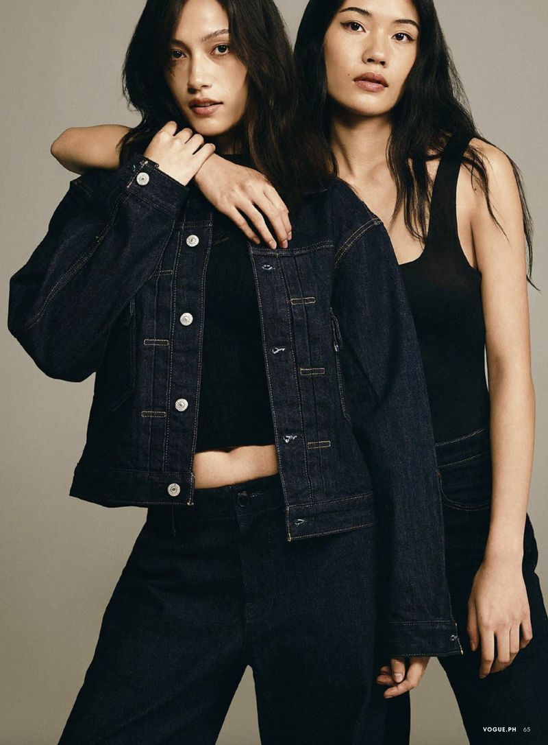 Two Asian women pose indoors wearing dark denim jacket and pants. One woman has her arm over the other's shoulder in a studio shot.