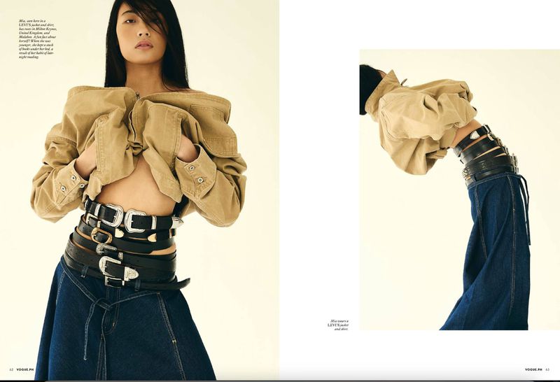 Editorial fashion shot of an Asian female model in a beige jacket and denim skirt, layered with multiple leather belts against a cream background.