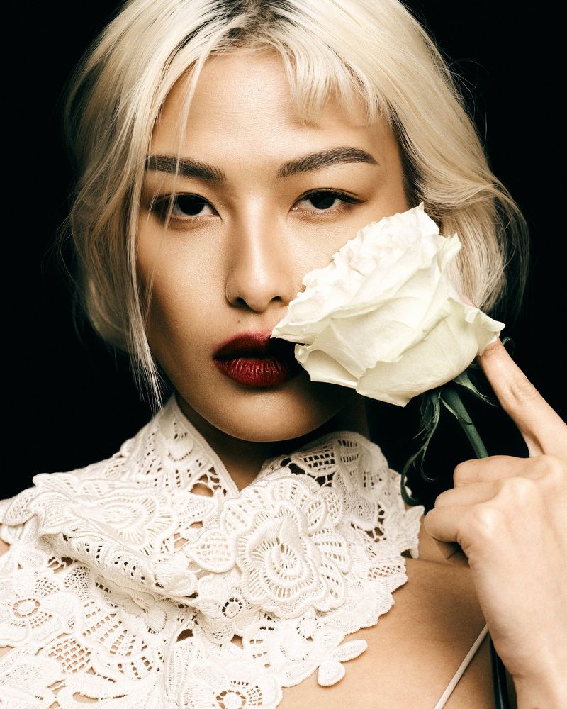 Close-up portrait of a woman with dyed blonde hair and dark red lipstick, posed with a white rose against a dark, high-contrast background.