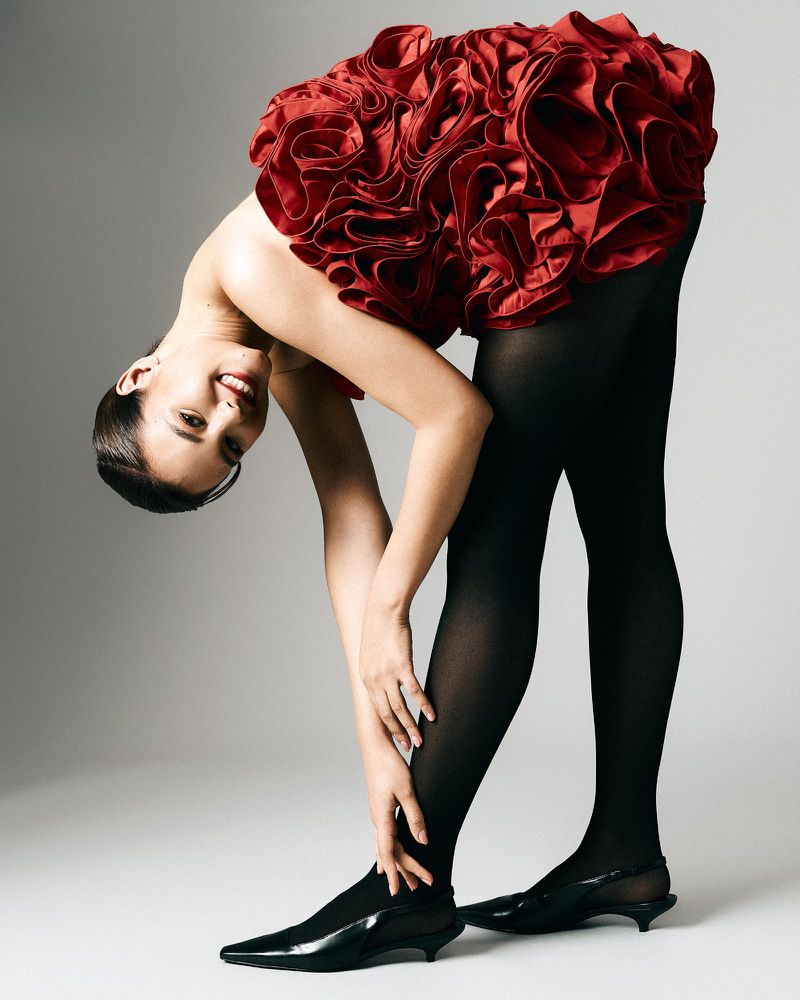 A woman in a red ruffled dress and black tights bends over with a smile in a studio shot, showcasing fashion and posing.