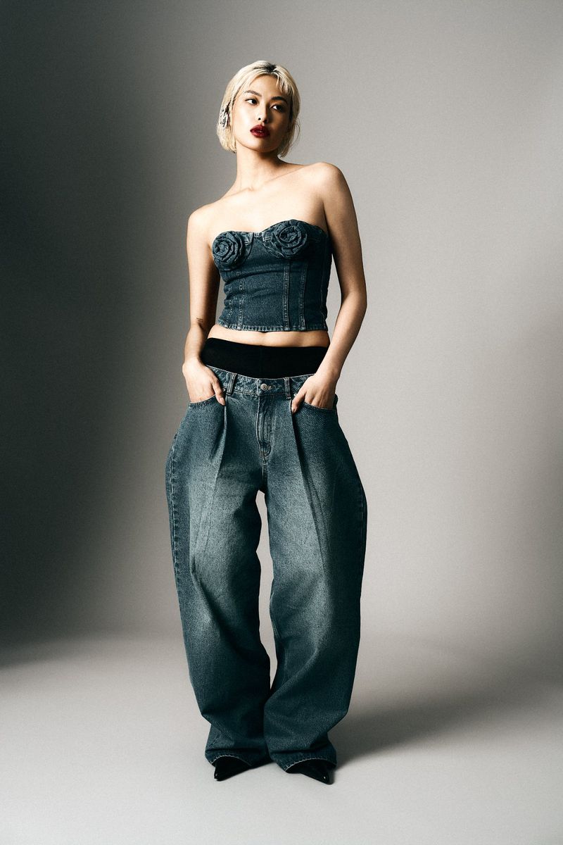 Woman in a denim tube top and oversized jeans stands in a photo studio with gray background, showcasing a fashion forward and unique style.