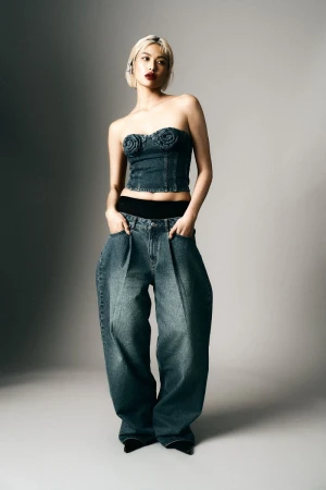 Woman in a denim tube top and oversized jeans stands in a photo studio with gray background, showcasing a fashion forward and unique style.