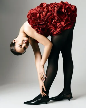 Model bending forward in a red rosette ruffle mini dress, black tights, and pointed black kitten heels on a seamless studio backdrop.