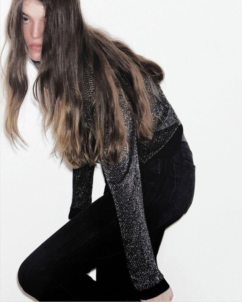 Fashion portrait of a young adult woman with long brown hair wearing a silver top and black jeans against a white background.