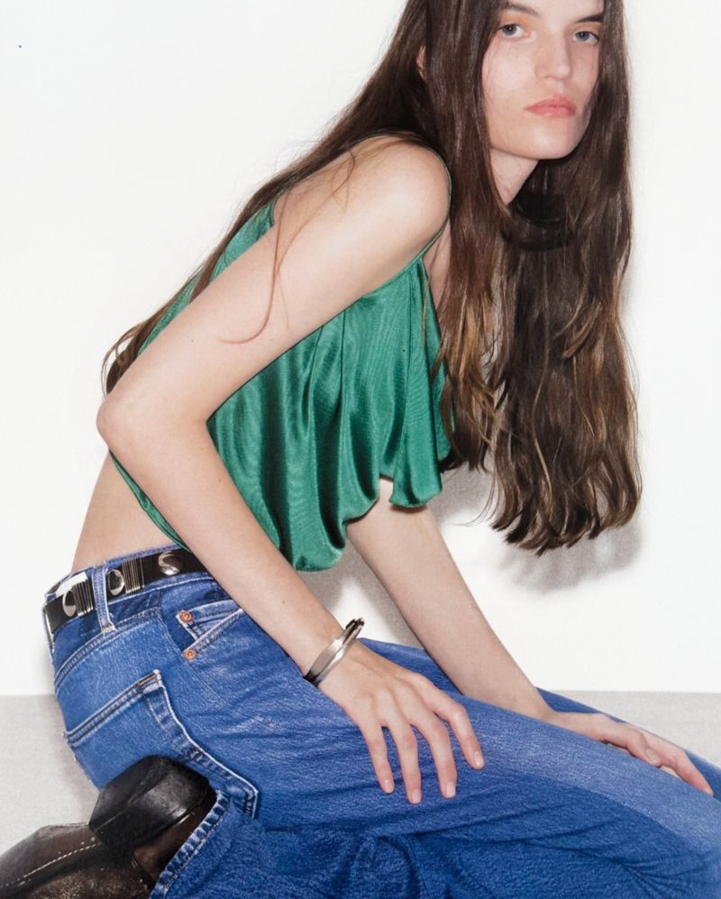 A slender model poses against a white background, wearing a shiny emerald green top, blue jeans with a black belt, and black shoes.
