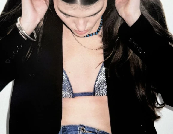 Young woman poses indoors wearing a sequined bikini top, blue jeans, and a black blazer, with dark hair and a light skin tone.