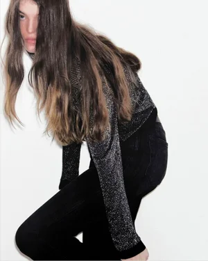 Model with long brown hair wearing a metallic sparkle top and black jeans against a white background