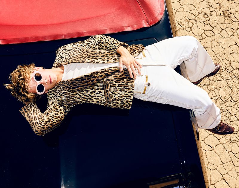 Man in animal-print jacket and white pants lies on a car in the desert sun, wearing sunglasses, exuding retro fashion vibes.