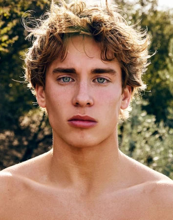 Close-up portrait of a fair-skinned young adult with blond wavy hair and blue eyes, posing outdoors with a blurred natural background.